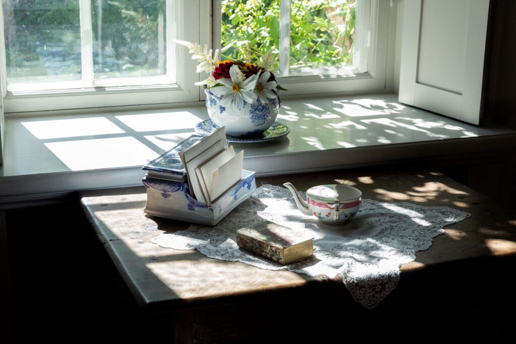 Ett solbelyst gammaldags skrivbord vid ett fönster med en spetsduk, en liten tekanna, en bok, och ett porslinsställ med brev och vykort. På fönsterbrädan står en blåvit porslinsskål med färska blommor.