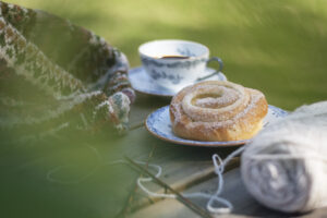 En bild på en kanelbulle på ett fat och en kopp kaffe, omgivet av garn och stickningar